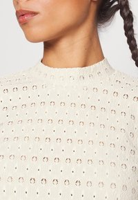 Gros plan sur une personne portant un haut blanc texturé avec des motifs circulaires en œillets et un col montant, les cheveux coiffés en tresse.