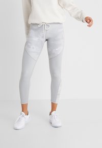 Person iført lysegrå camo-mønstrede tights med snorlukning i taljen og hvide sneakers foran en ensfarvet hvid baggrund.