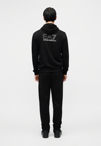 Μαύρο φούτερ με κουκούλα και παντελόνι σετ από απαλή ύφανση. Το φούτερ έχει λογότυπο στην πλάτη. Το σχέδιο είναι σε άνετη γραμμή με ριμπ τελειώματα στα μανίκια.