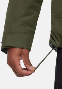 Grøn vandtæt jakke med en snørelukning ved håndleddet, glat tekstur og elastisk manchet detaljer.