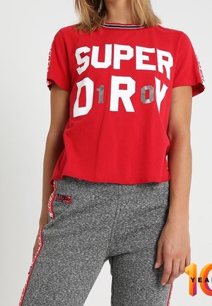 Red short-sleeve T-shirt with large white "SUPERDRY" print. Paired with gray sweatpants featuring side stripes and a logo patch on the pocket.