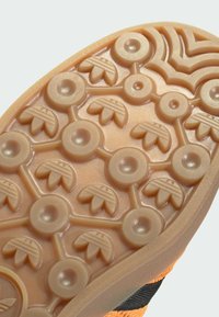 Gumeni potplat tenisica u smeškastoj boji s teksturiranim uzorkom, koji sadrži kružne i oblike nalik na lišće za prianjanje i trakciju. Vidljivi crni akcenti.