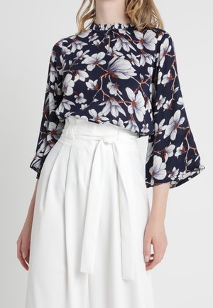 Blouse bleu marine à imprimé floral blanc avec manches larges, associée à un pantalon blanc taille haute avec ceinture en tissu nouée.
