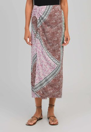 Femme portant une jupe midi portefeuille avec des motifs floraux roses et marron et des sandales noires à brides à la cheville, debout devant un fond gris.