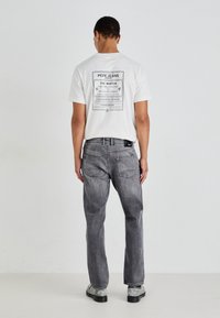 Λευκό μπλουζάκι με μαύρο γραφικό σχέδιο στην πλάτη; συνδυασμένο με γκρίζα τζιν παντελόνια με ίσια γραμμή και πίσω τσέπες. Μαύρα υποδήματα.