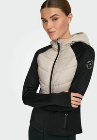 Lahka jakna s kapuco, z vstavkom iz bež prešite tkanine na zgornjem delu in črnim rokavom. Ima zadrgo po celotni dolžini, raztegljivo tkanino ter logotip na rokavu.