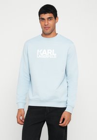 Svetlomodrá mikina s rebrovanými manžetami a lemom, s bielou potlačou loga "KARL LAGERFELD" na hrudi. Mäkká textúra látky.