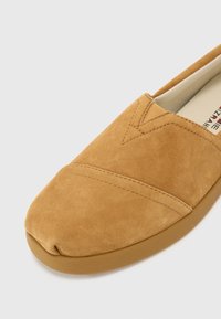 Smeđi nubuk cipelama bez vezivanja s zaobljenim vrhom, s detaljima ukrštenog šavljenja i gumeni potplat za bolj grip. Glatka tekstura kroz cijelu cipelu.