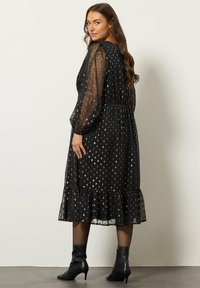 Musta mekko, jossa on läpinäkyvät puffihihat, pisteitä kullanvärisiä koristeita, kerroksellinen helma ja mukautettu vyötärö. Yhdistetty mustiin nilkkureihin.