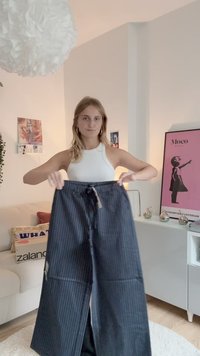 Pantalons larges à rayures noires avec une taille à cordon de serrage, présentant une texture lisse et un subtil éclat, tenus dans un environnement intérieur lumineux.
