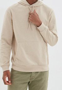 Felpa beige in cotone con tasca frontale e cappuccio con stringhe. Presenta polsini e orlo a costine. Le mani del modello sono posizionate all'altezza del collo.