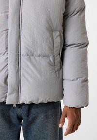 Lichtgrijze pufferjack met een gladde textuur, brede gebreide secties en een zijzak, gecombineerd met blauwe spijkerbroeken.