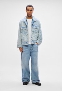 Man standing with hand in pocket wearing a textured light blue jacket, white shirt, wide-leg blue jeans, and black shoes on white background.