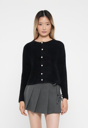 Cardigan noir en peluche avec un col rond, comportant six boutons en perles sur le devant. Associé à une jupe grise plissée.