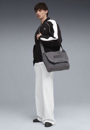 Ung person iført sort og hvid Puma-jakke, hvide brede bukser, sorte sko, der holder en grå Puma skuldertaske mod en neutral baggrund.