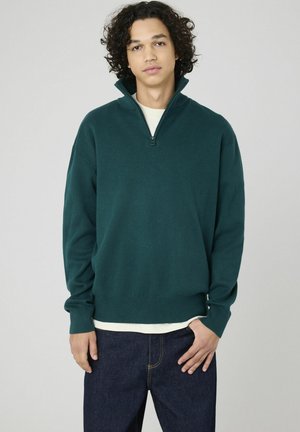 Sweatshirt à col zippé vert en tissu doux, avec des manches longues et un ourlet côtelé, associé à un jean bleu foncé.