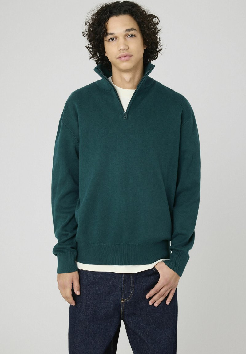 Sweatshirt à col zippé vert en tissu doux, avec des manches longues et un ourlet côtelé, associé à un jean bleu foncé.