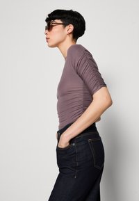 Upravený mauve top s nařasenými rukávy, kombinovaný s tmavými džíny. Tento outfit disponuje hladkou texturou a jednoduchými liniemi, které jsou zvýrazněny kapsami.