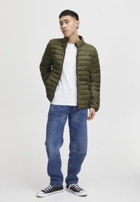 Olive green puffer jacket with quilted texture, stand-up collar, and zipper; worn over a white shirt and blue jeans, paired with black sneakers.