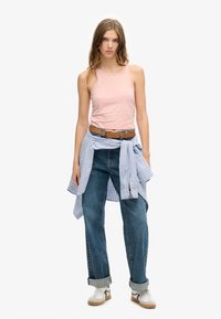 Lichtroze tanktop, blauw gestreepte shirt geknoopt in de taille, losse blauwe spijkerbroek en witte sneakers met bruine accenten.