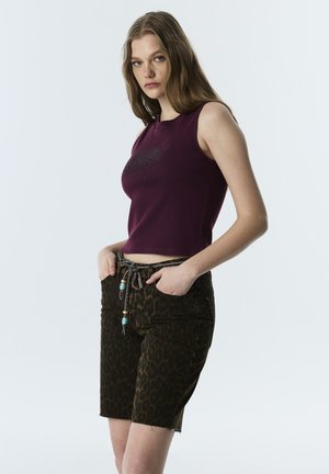 Junge Frau mit langen Haaren, die ein ärmelloses lila Oberteil und knielange Shorts mit Leopardenmuster und Perlengürtel trägt, Hände in den Taschen.