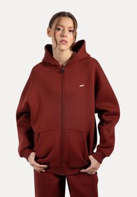 Burgundowa bluza z kapturem na zamek, wykonana z miękkiego materiału, z dużą kieszenią z przodu i ściągaczami na rękawach. Posiada kaptur i mały detal z logo.