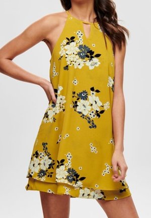 Ärmelloses senfgelbes Kleid mit weißen und schwarzen Blumenmustern, Neckholder-Ausschnitt und mehrlagigem Saum getragen von einer stehenden Frau.