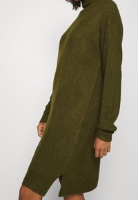 En person som bär en olivgrön stickad turtleneck klänning med långa ärmar och sidoklyfta, sedd från mitten av låret till axeln.