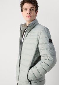 Light gray puffer jacket with a high collar, zip closure, and horizontal quilted pattern. Features black logo patch on sleeve.