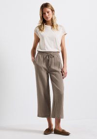 Lichtbeige top met kantdetail op de mouwen; losse, gestructureerde bruine broek met een elastische tailleband en zijzakken; platte bruine schoenen.