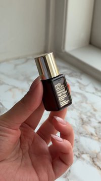Main tenant un petit flacon brun foncé Estée Lauder Advanced Night Repair avec un bouchon doré au-dessus d'une surface en marbre blanc proche d'une fenêtre.
