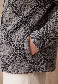 Hallikas ja tumesinine mustriga jakk pehme tekstuuriga, millel on küljetasku ja vabaaja disain. Kangas on sümmeetrilise lillemustriga.