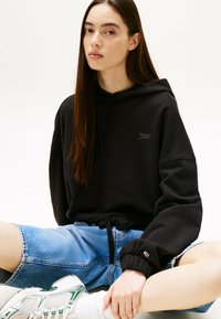 Sudadera negra con capucha y cordón, con un logo sutil, combinada con unos pantalones cortos de mezclilla azul claros y zapatillas deportivas con detalles verdes.