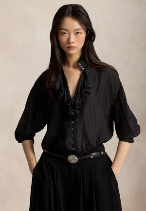 Femme aux longs cheveux foncés portant un chemisier noir à volants avec des boutons et une jupe noire avec une ceinture décorative à boucle argentée, mains dans les poches.