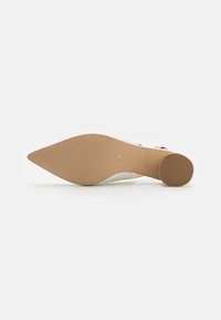 Chaussure à talon blanc avec un bout pointu, extérieur lisse, détail de bride transparente et semelle beige texturée présentant un léger motif en rainures.