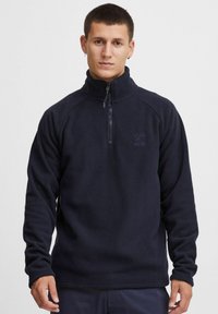Marinblå fleecepullover med hög krage, halv dragkedja och långa ärmar. Har en liten logotyp på bröstet och en mjuk, texturerad yta.