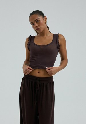 Femme portant un haut court sans manches marron foncé et un pantalon ample, posant devant un fond clair uni.