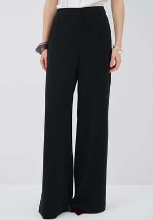 Pantalones anchos de talle alto y color negro, confeccionados en una tela suave. Presentan un diseño frontal limpio y detalles de costura sutiles, combinados con una pulsera.