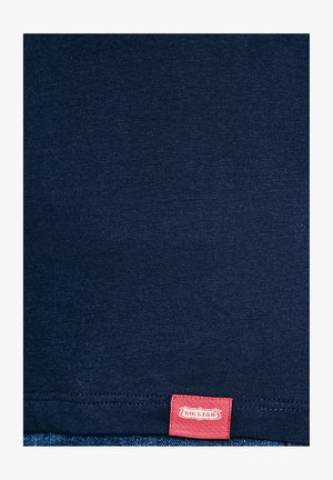 Zbliżenie na granatowy materiał z widocznym przeszyciem i małą czerwoną prostokątną metką z napisem "BIG STAR" na dolnym brzegu.