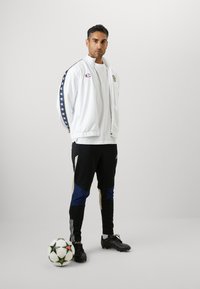 Witte sportieve zip-up jas met blauwe en rode strepen, zwarte en blauwe trainingsbroek en zwarte voetbalschoenen. Een voetbal bij zijn voeten.
