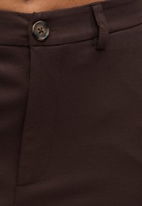 Καφέ υφασμάτινο παντελόνι με ένα κουμπί στη μέση, επίπεδη εμπρόσθια επιφάνεια και ραμμένες λεπτομέρειες. Το υλικό φαίνεται λείο και δομημένο.