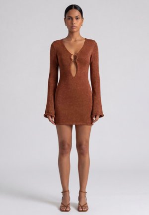 Donna che indossa un mini abito marrone in maglia a maniche lunghe con un'apertura a goccia sul davanti e dettaglio ad anello, abbinato a sandali marroni con cinturini e tacco alto.