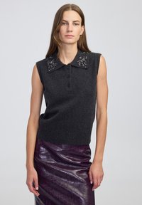 Kobieta w bezrękawniku w ciemnoszarym kolorze z ozdobnym ko�łnierzykiem oraz błyszczącej fioletowej, teksturowanej spódnicy, stojąca na tle jasnym.