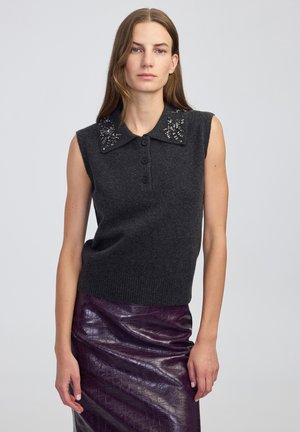 Femme portant un haut en tricot sans manches gris foncé avec un col orné et une jupe texturée en violet brillant, se tenant devant un fond clair.