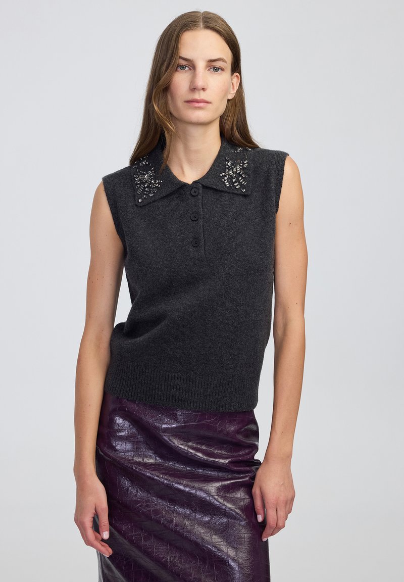 Kobieta w bezrękawniku w ciemnoszarym kolorze z ozdobnym kołnierzykiem oraz błyszczącej fioletowej, teksturowanej spódnicy, stojąca na tle jasnym.