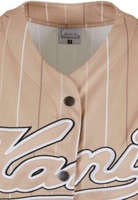 Beige Baseball-Trikot mit vertikalen weißen Streifen, einem V-Ausschnitt, zwei Druckknöpfen und besticktem Schriftzug in Schwarz und Weiß.