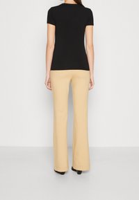 Femmes portant un haut noir à manches courtes et un pantalon beige à jambes larges, debout de dos devant un fond clair uni.