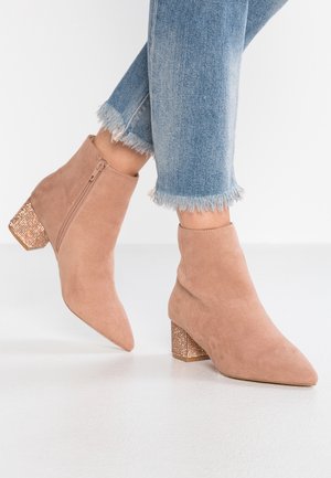 Beige ruskind ankelstøvler med sidelukning med lynlås og glitrende blokhæle, båret med slidte blå jeans mod hvidt gulv og væg.