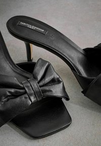 Sandales à talons hauts noires en cuir, avec un détail de nœud noué. Le design comprend un bout carré et un talon élancé pour une élégance supplémentaire.