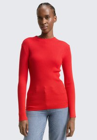 Roter, gerippter Langarmpullover mit Rundhalsausschnitt, der eine figurbetonte Silhouette und kontrastierende Bündchen hat. Kombiniert mit blauen Jeans.
